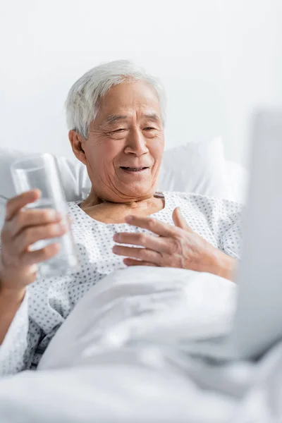 Sorrindo asiático paciente segurando vidro de água perto desfocado laptop no hospital cama — Fotografia de Stock