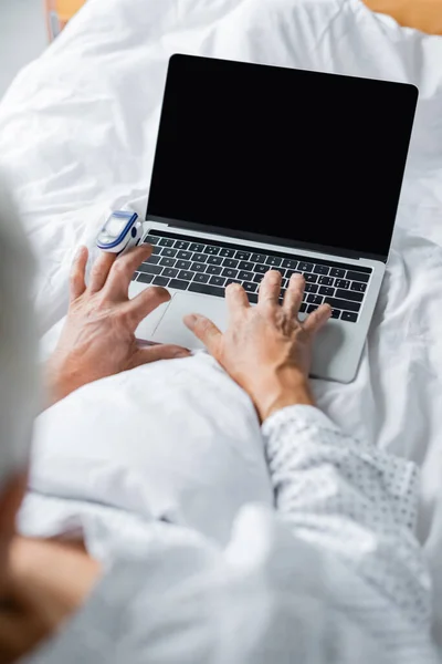 Paciente desfocado com oxímetro de pulso usando laptop com tela em branco na cama — Fotografia de Stock