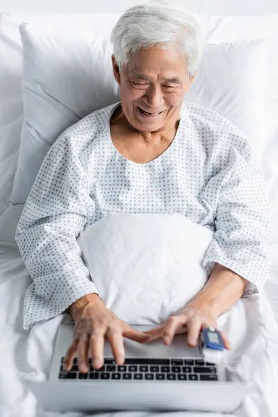 Positive asian patient with pulse oximeter using blurred laptop on bed in clinic — Stock Photo