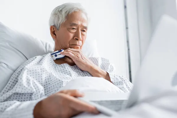 Elderly asian patient with pulse oximeter holding blurred laptop in hospital ward — Stock Photo