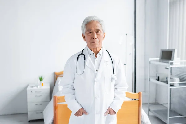 Asian doctor in white coat looking at camera in hospital ward — Stock Photo