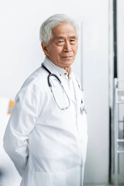 Personnes âgées asiatique médecin en blanc manteau regarder caméra à l'hôpital — Photo de stock