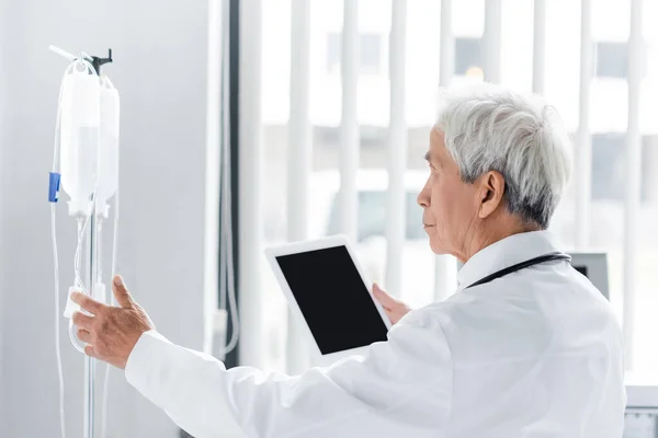 Vista lateral del médico de edad avanzada asiático con tableta digital mirando soporte de terapia intravenosa en la clínica - foto de stock