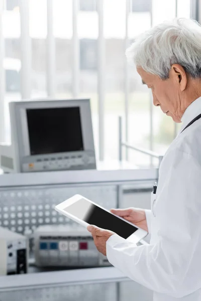 Пожилой азиатский врач с цифровым планшетом рядом с размытым медицинским оборудованием в больнице — стоковое фото