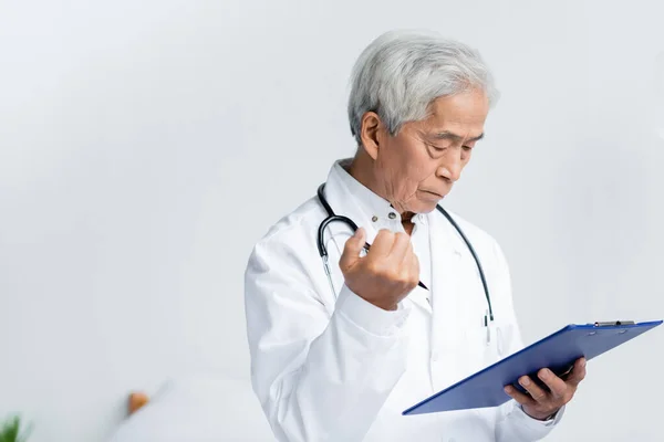Elderly asian doctor with pen and clipboard standing in hospital — Stock Photo