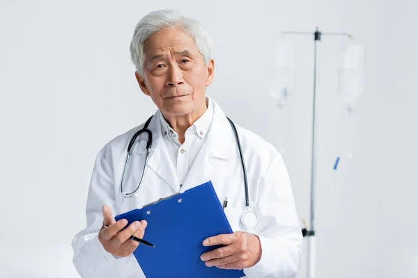 Senior asiatische Arzt mit Stift und Klemmbrett Blick auf Kamera im Krankenhaus — Stockfoto