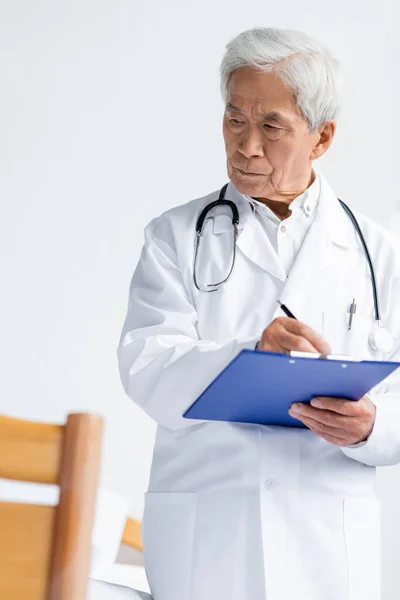 Ancianos asiático médico en blanco abrigo escritura en portapapeles en hospital sala - foto de stock