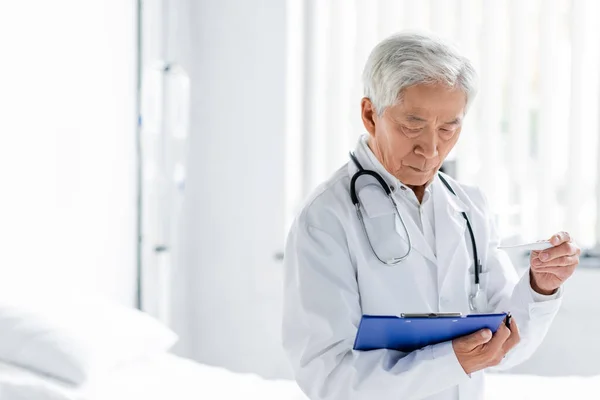Senior asian doctor holding thermometer and clipboard in hospital ward — Stock Photo