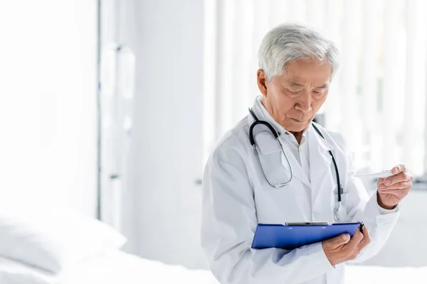 Senior asiático médico celebración termómetro y portapapeles en hospital sala - foto de stock