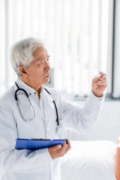 Asiatischer Arzt mit Klemmbrett schaut auf Thermometer in Klinik — Stockfoto