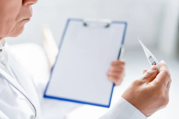 Cropped view of doctor holding thermometer and blurred clipboard in clinic — Stock Photo