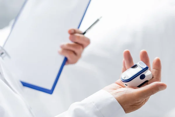 Ausgeschnittene Ansicht des Pulsoximeters und verschwommenes Clipboard in der Hand des Arztes in der Klinik — Stockfoto