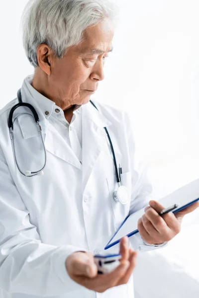 Asiatischer Arzt blickt auf Klemmbrett und hält verschwommenes Pulsoximeter in Klinik — Stockfoto