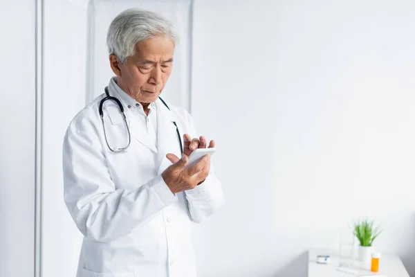 Senior asian doctor in white coat using smartphone in hospital — Stock Photo