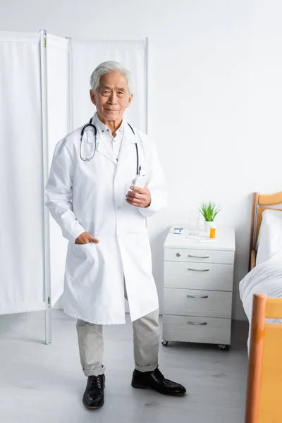 Médecin asiatique senior tenant smartphone près du lit dans la salle d'hôpital — Photo de stock