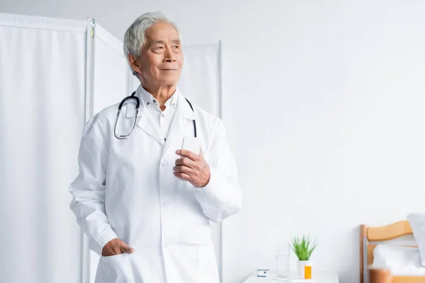 Elderly asian doctor with cellphone standing in hospital ward — Stock Photo