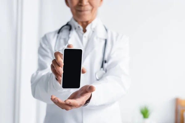 Обрезанный вид смартфона в руках размытого врача в клинике — стоковое фото