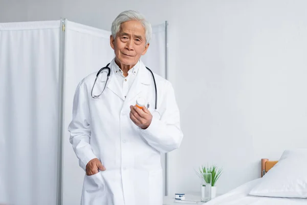 Médecin asiatique senior tenant des pilules et regardant la caméra dans la clinique — Photo de stock