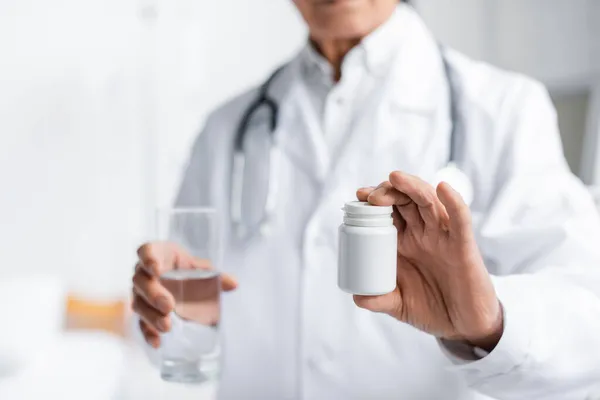 Vista recortada del médico borroso sosteniendo frasco con pastillas y vaso de agua en la clínica - foto de stock