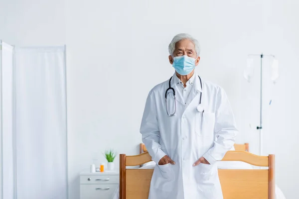 Senior asiático médico en médico máscara mirando a cámara en hospital sala - foto de stock