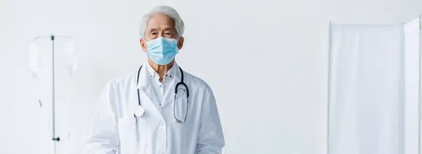 Idoso asiático médico em máscara médica e casaco branco olhando para a câmera na clínica, banner — Fotografia de Stock
