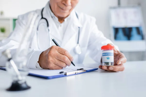 Vista recortada del médico escribiendo en el portapapeles y sosteniendo cannabis medicinal en la clínica - foto de stock