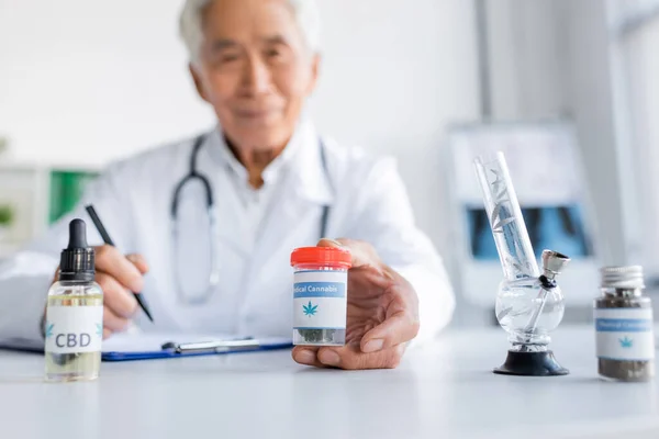 Blurred asian doctor holding medical cannabis near bong and cbd oil in clinic — Stock Photo