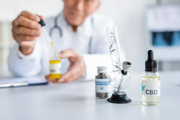 Cropped view of cbd oil and glass bong near blurred doctor in hospital — Stock Photo