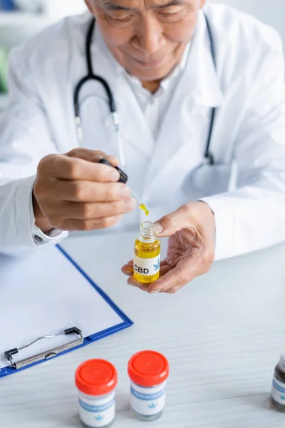Médico asiático sosteniendo aceite de cbd cerca de cannabis medicinal y portapapeles en la clínica - foto de stock
