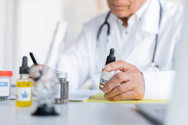 Vista recortada del doctor borroso sosteniendo aceite de cbd cerca de bong y cannabis medicinal en la clínica - foto de stock