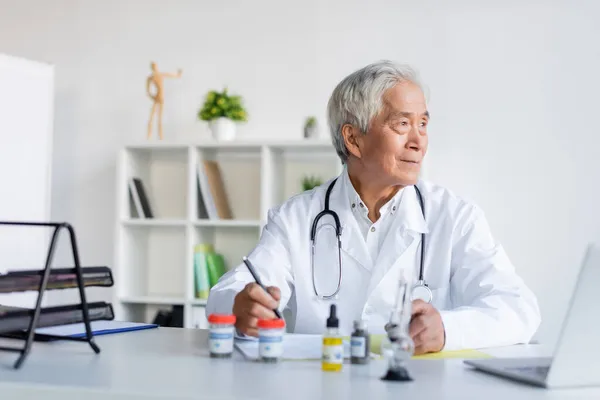 Médico asiático sosteniendo pluma cerca de aceite de cbd, pipa y portátil en la clínica - foto de stock