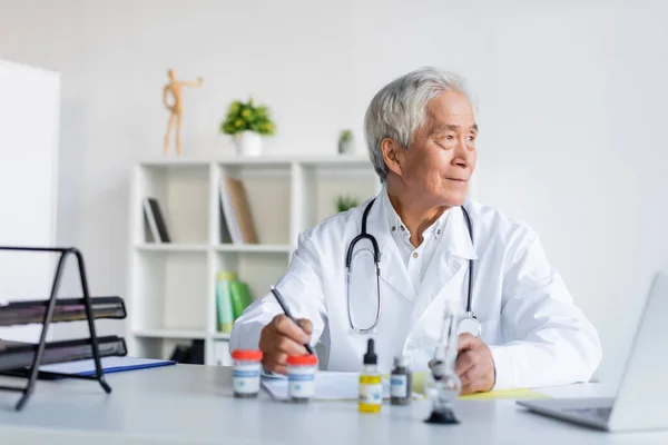 Asiatico medico holding penna vicino cbd olio, bong e laptop in clinica — Foto stock