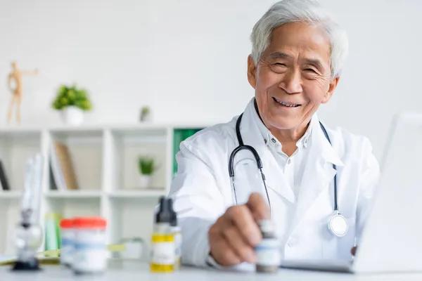 Smiling asian doctor looking at camera near medical cannabis and laptop in clinic — Stock Photo
