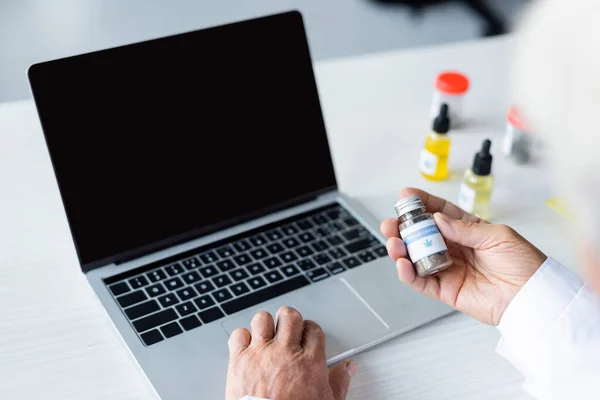 Vista cortada do médico segurando cannabis medicinal perto de laptop e óleo de CDB — Fotografia de Stock