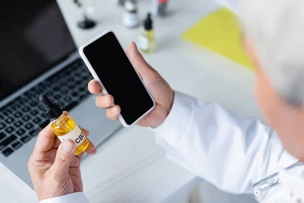 Medico offuscato che tiene smartphone e olio cbd vicino al computer portatile in ospedale — Foto stock