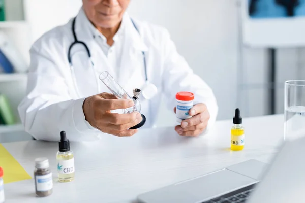 Cropped view of blurred doctor holding bong and medical cannabis near laptop and cbd oil — Stock Photo