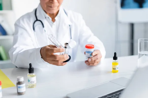 Vista cortada de médico desfocado segurando bong e cannabis medicinal perto de laptop e óleo de cbd — Fotografia de Stock