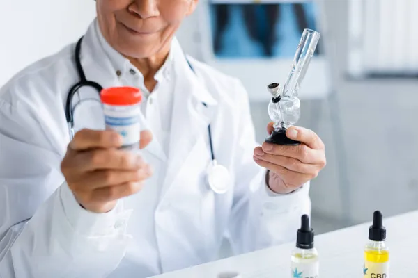 Vista cortada do médico segurando bong e cannabis medicinal perto de óleo de cbd na clínica — Fotografia de Stock