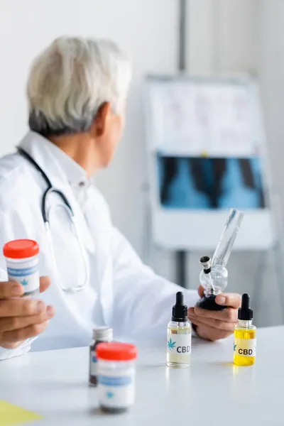 Senior doctor holding glass bong and medical cannabis near cbd oil in hospital — Stock Photo