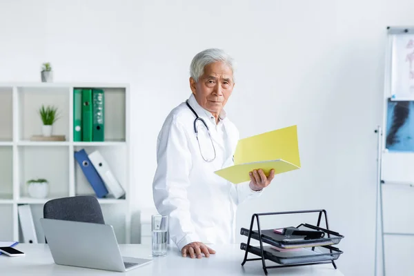Senior asian doctor holding paper folder near devices and glass of water in clinic — Stock Photo