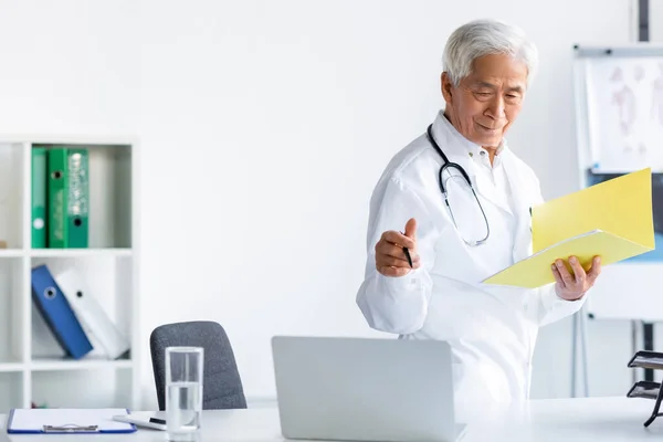 Médecin principal avec stylo et dossier papier regardant l'ordinateur près de l'eau et presse-papiers dans la clinique — Photo de stock