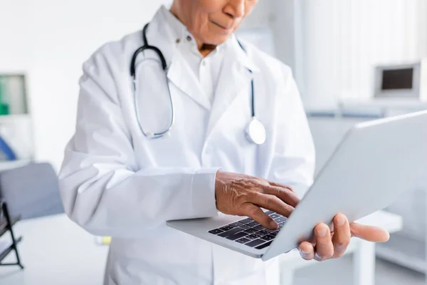 Vista cortada de computador portátil em mãos de médico sênior em casaco branco na clínica — Fotografia de Stock