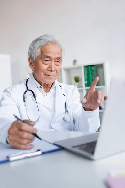 Senior médico asiático con portapapeles mirando portátil durante la consulta en línea en el ordenador portátil en la clínica - foto de stock