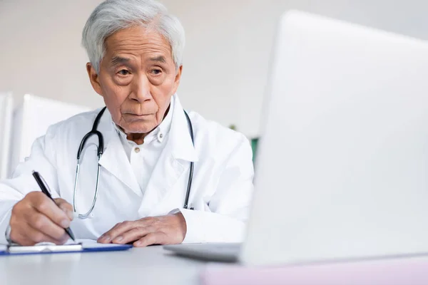 Elderly asian doctor writing on clipboard near blurred laptop in hospital — Stock Photo