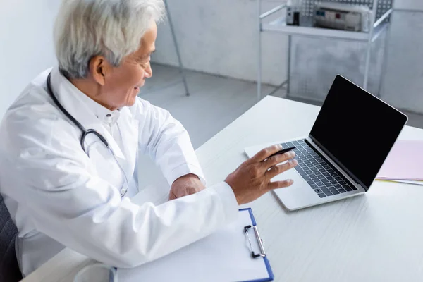 Vue latérale du médecin asiatique souriant avec stylo à l'aide d'un ordinateur portable près du presse-papiers dans la clinique — Photo de stock