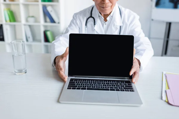 Vista ritagliata del medico anziano che tiene il computer portatile con schermo vuoto vicino a vetro d'acqua in clinica — Foto stock