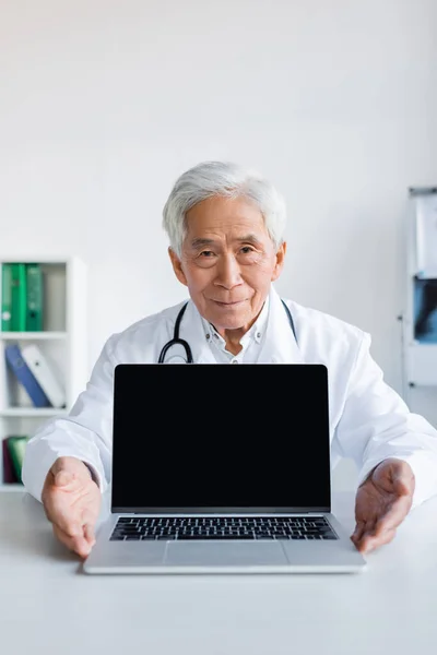 Anziano asiatico medico guardando fotocamera vicino laptop con vuoto schermo in ospedale — Foto stock