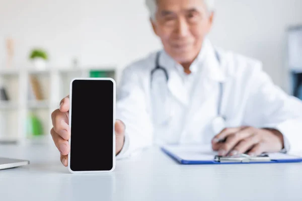 Blurred asiático médico celebración de teléfono inteligente con pantalla en blanco cerca de portapapeles en el hospital - foto de stock