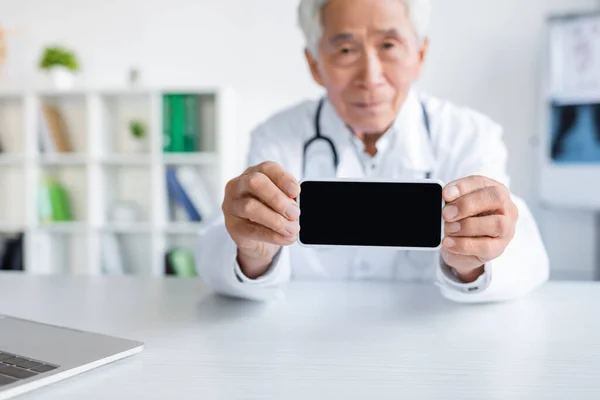 Smartphone con schermo bianco nelle mani del medico asiatico offuscato in ospedale — Foto stock