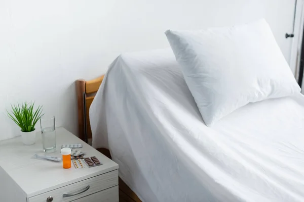 Pills and glass of water on bedside table near bed in hospital ward — Stock Photo